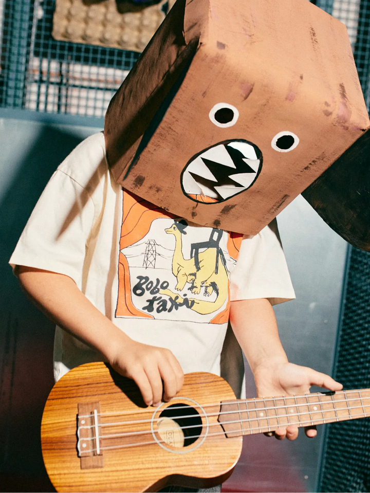 A kid wearing a large brown cardboard dog-head mask with cartoon eyes and a mouth, playing a wooden ukulele. The person is wearing a white graphic t-shirt with a cartoon dinosaur and Japanese text.